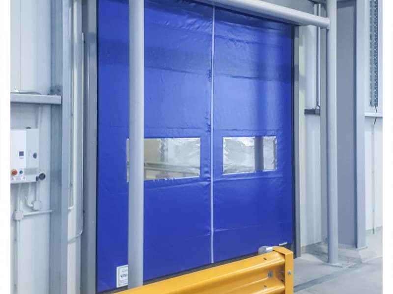 Blue high-speed roll door with vision panels and yellow safety barrier.